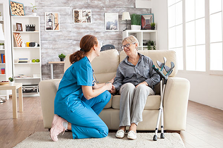 A female healthcare worker in scrubs talking with an elderly woman in her home.