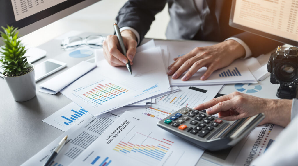 Two tax professionals collaborate on year-end tax strategy planning, with one writing on financial documents and the other using a calculator over a desk filled with charts and graphs.