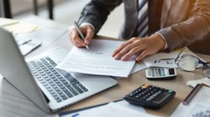 A business owner in a suit signing an LLC formation document at a desk with a laptop and calculator.